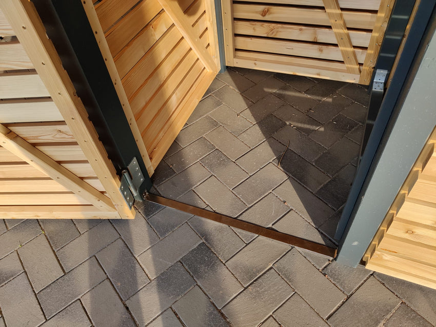 Innenansicht der Mülltonnenbox KLARE LINIE mit geöffneter Tür und Bodenstrebe auf Pflasterfläche, stabile Holz- und Metallkonstruktion.